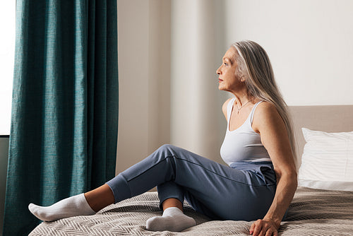 Loneliness aged woman sitting on a bed looking at window at home