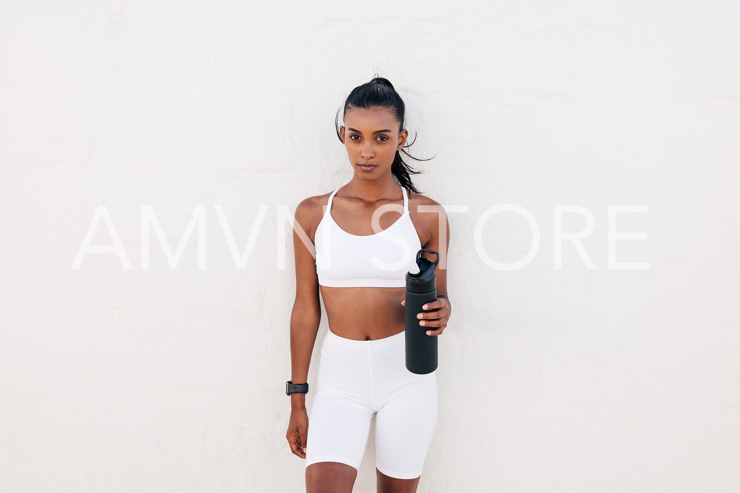 Slim woman in white fitness attire holding bottle leaning wall looking at the camera