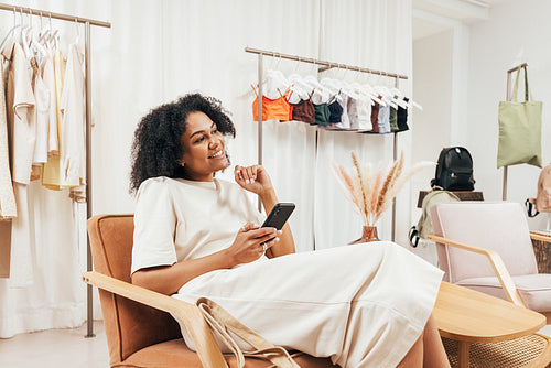 Side view of a stylish customer holding mobile phone while relaxing on armchair during shopping in a boutique