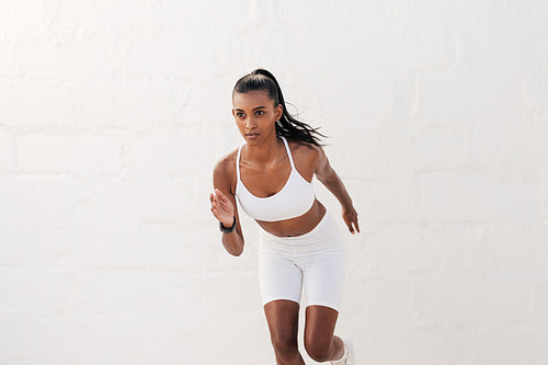 Young slim female athlete running outdoors against a white wall. Fitness influencer in white sportswear sprinting.