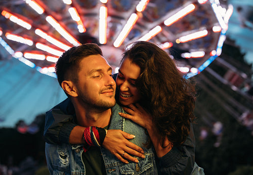 Smiling man piggybacks his happy girlfriend at the festival. Young couple having fun at night in an amusement park.