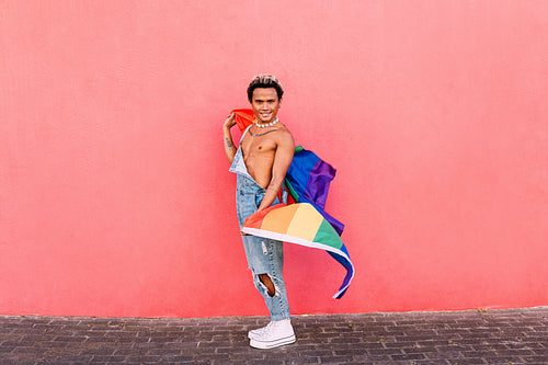 Young confident guy holding a rainbow LGBT flag and looking at camera at pink wall