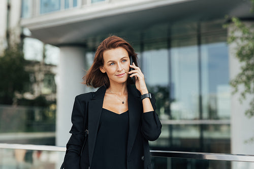 Confident businesswoman with ginger hair making a phone call while walking outdoors against office building