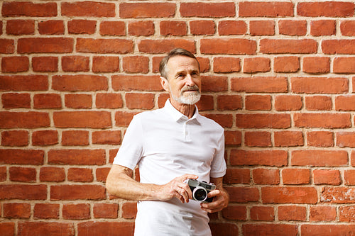 Bearded senior man standing with a film camera at a brick wall