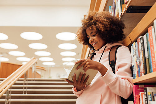 Smiling girl reading a book while leaning a bookshelf in library