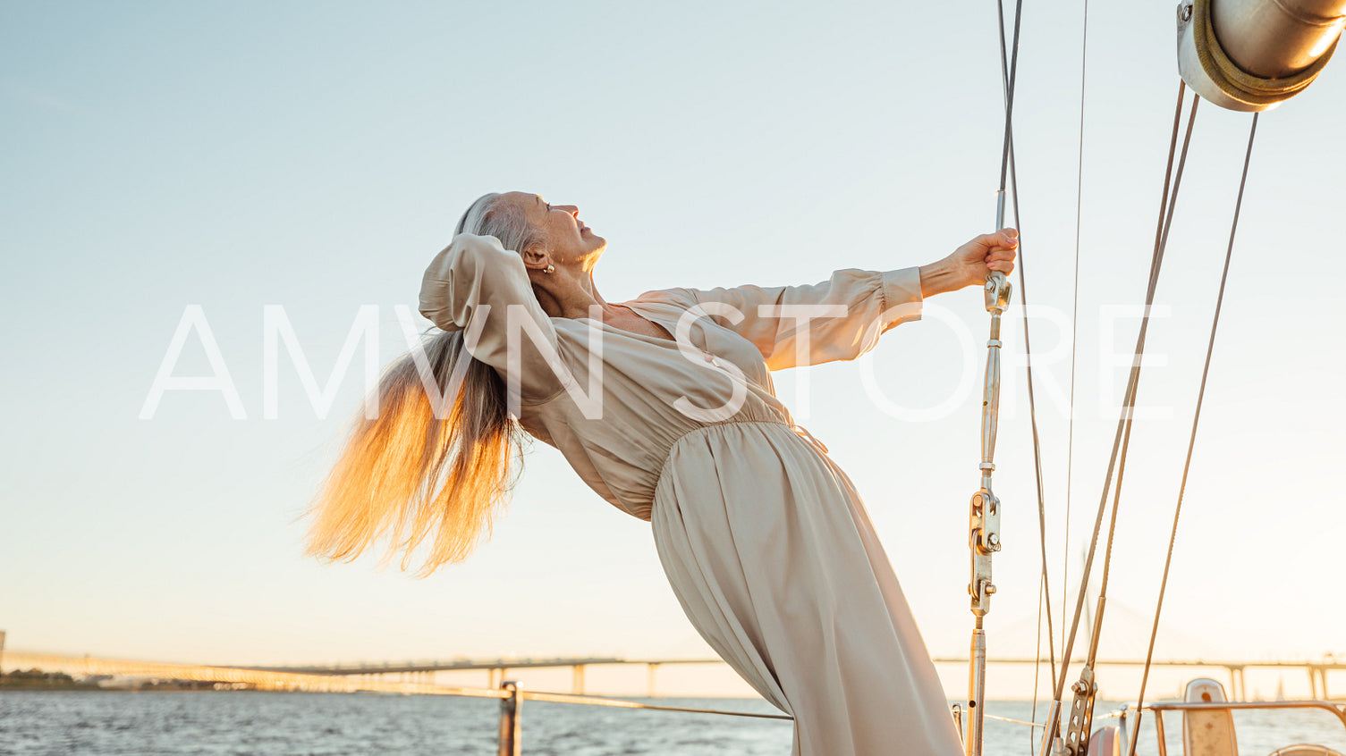 Side view of a mature woman with long grey hair holding a rope on a private yacht and enjoying sunset	