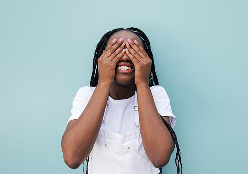 Smiling female with braids hiding her face with palms while standing at a blue wall outdoors