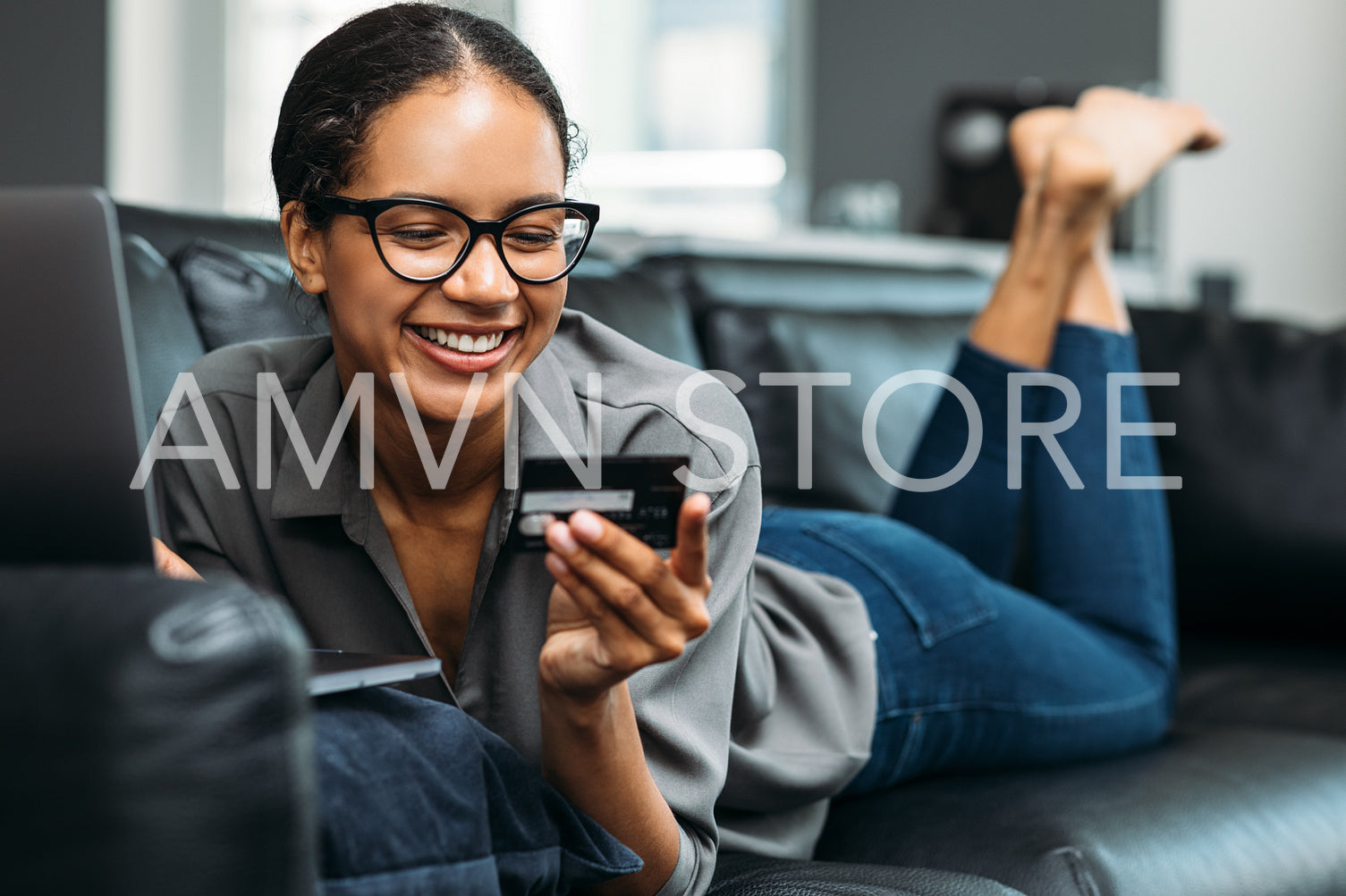 Happy young woman at home shopping online. Smiling female holding a credit card while lying on a sofa in a living room.	