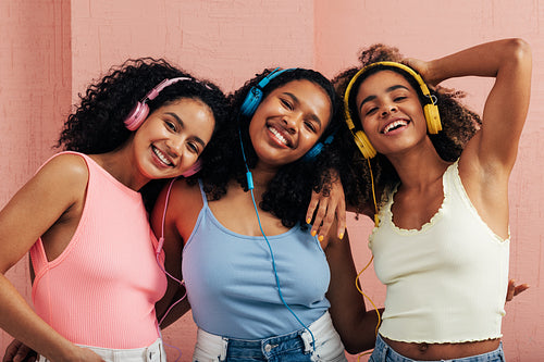 Portrait of three young women wearing headphones enjoying music against a pink wall