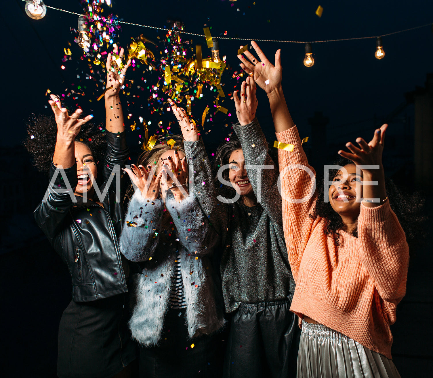 Four laughing females throw confetti in the air at night. Diverse women celebrating together.