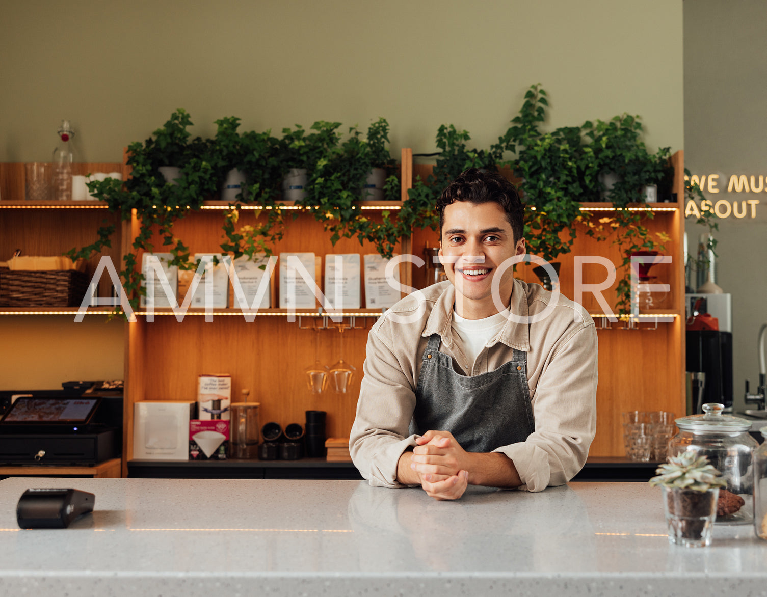 Smiling coffee shop owner in apron leaning a counter. Middle eastern bartender looking at camera.