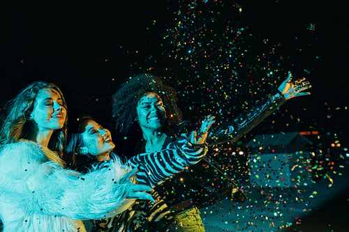 Three happy girls throwing confetti while standing on the roof under neon lights