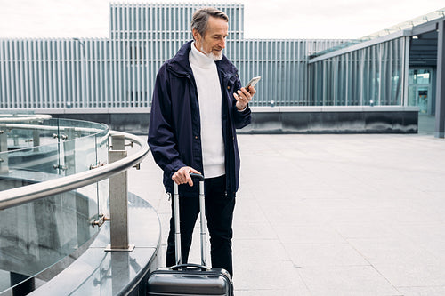 Senior businessman standing outdoors with suitcase and looking on smartphone