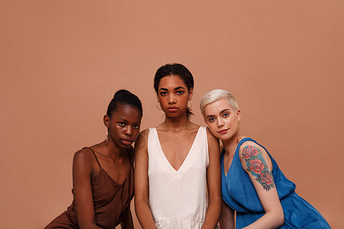 Three beautiful women with different skin color looking at camera. Young females in dresses sitting on brown backdrop.