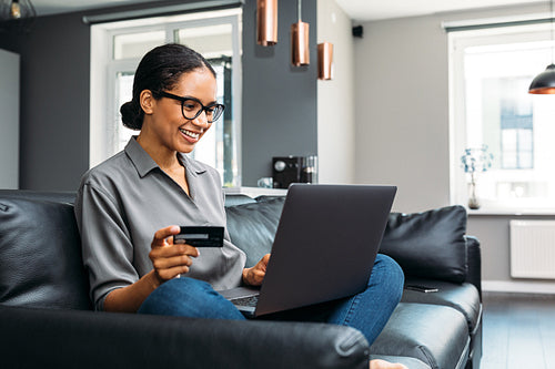 Happy woman sitting on a sofa in the living room holding a credit card with a laptop on her laps