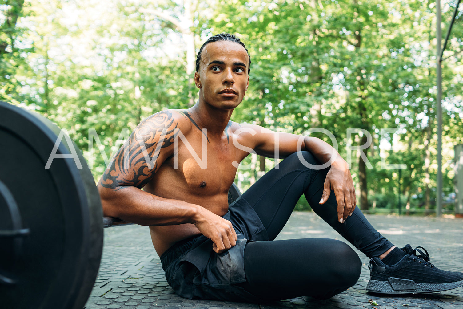 Muscular athlete sitting on sports ground with hand on barbell looking away	