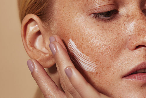 Close up highly detailed shot of a young woman applying cream on her freckled skin. Cropped shot of woman applying moisturizer on cheek with fingers.