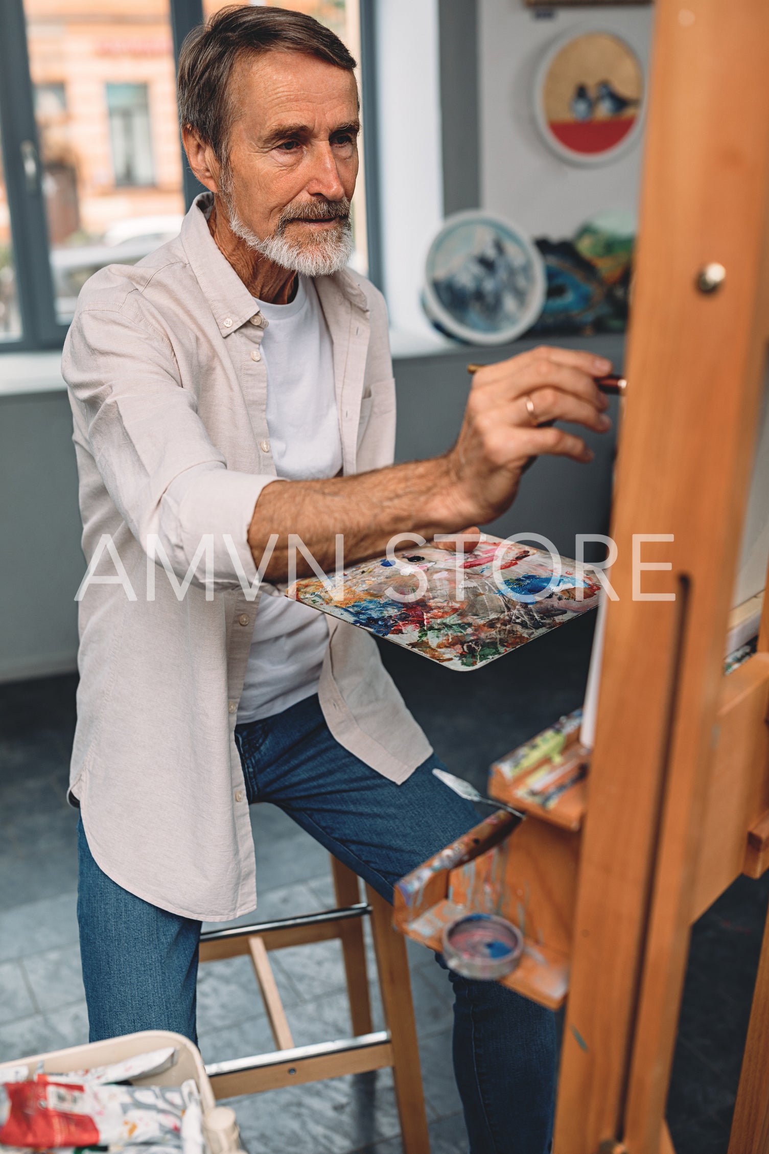 Bearded artist sitting in front of easel and painting abstract picture in studio	