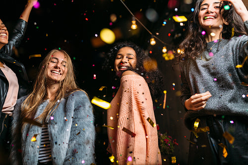 Female friends dancing under confetti at night, looking at camera