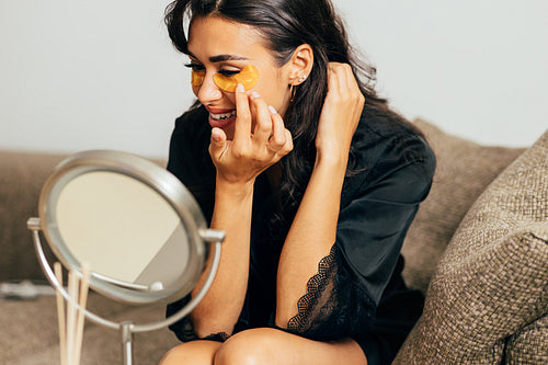 Young woman in sating robe sitting on a couch in front of a mirror applying an eye patch