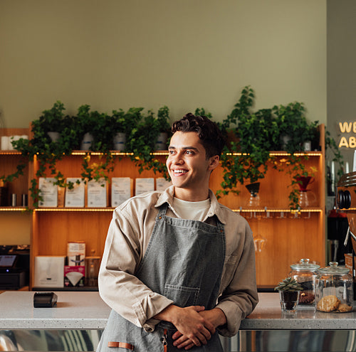 Portrait of a handsome coffee shop owner looking away. Young male in an apron leaning counter.