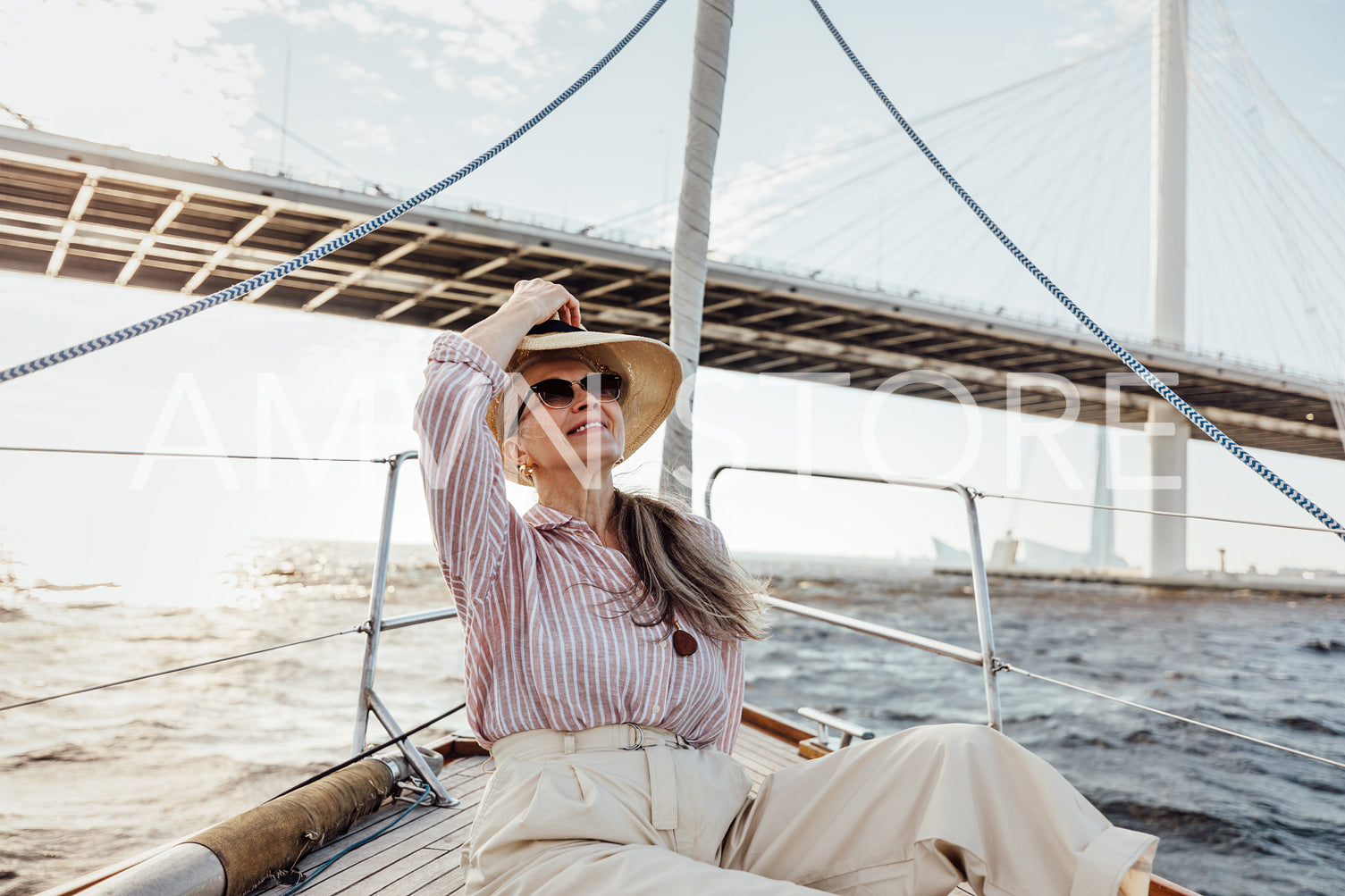 Mature woman in sunglasses puts on hat sitting on a sailboat at sunny day	