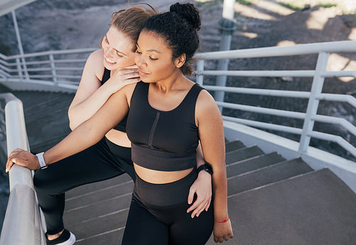 Two female friends in black fitness attire standing together on stairs. Two diverse athletes relax after exercise.