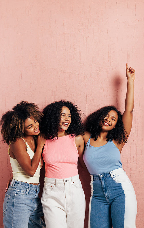 Three beautiful female friends in bright casuals having fun while leaning pink wall