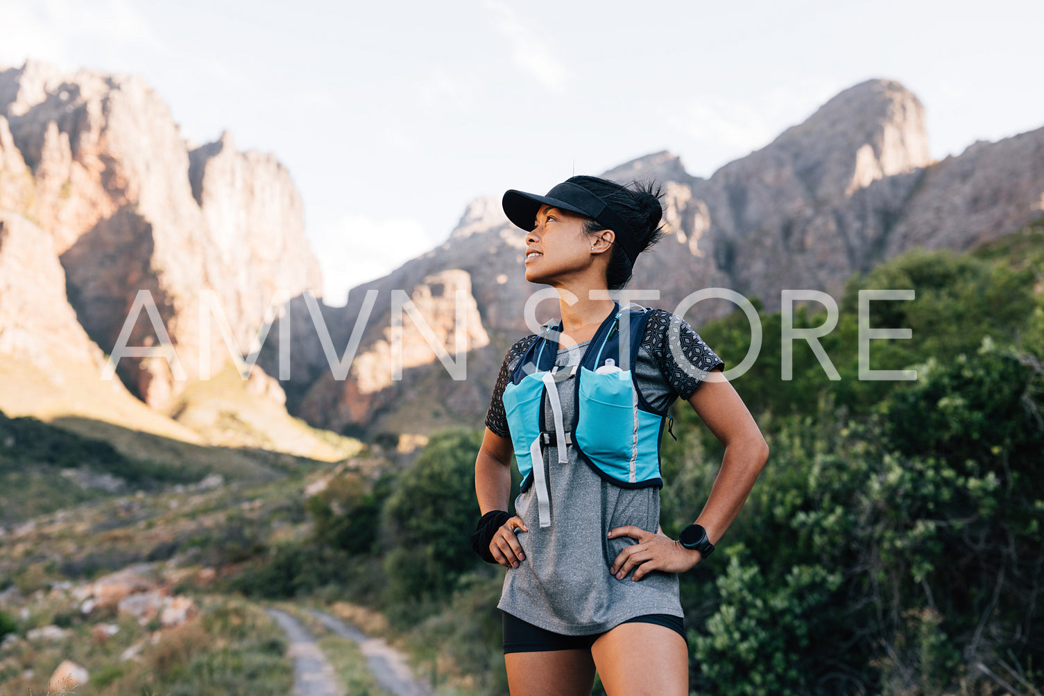 Trail runner in cap relaxing in valley. Sportswoman looking away and smiling preparing for hike.