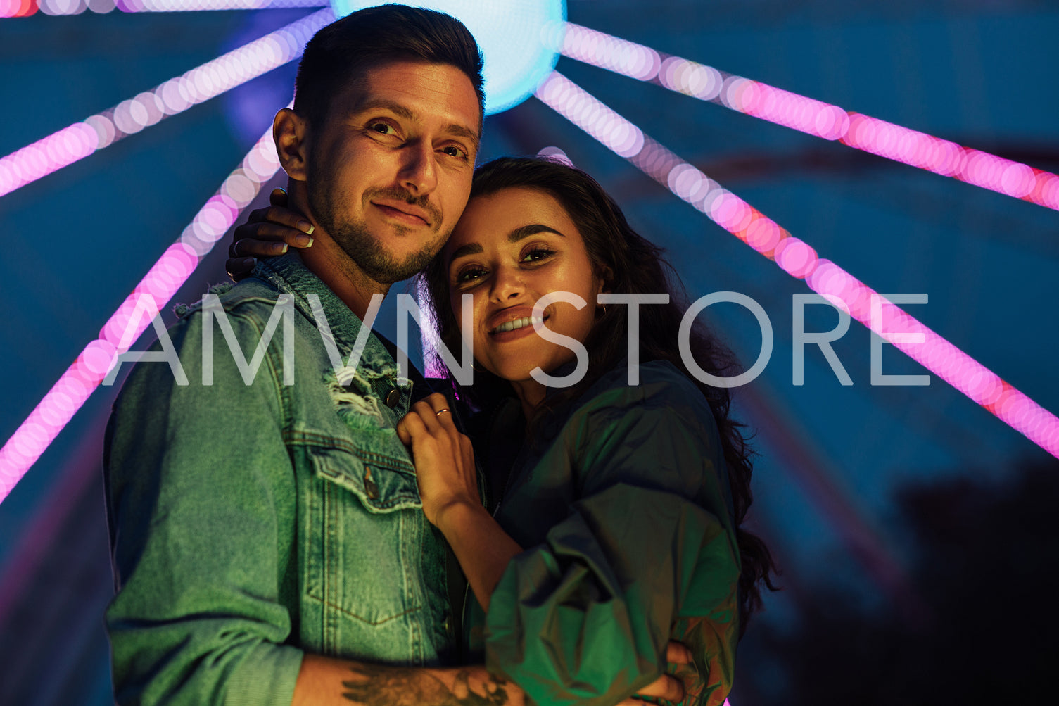 Young lovely couple hugging at night standing against the Ferris wheel. Girlfriend and boyfriend are standing together in an amusement park during the festival.