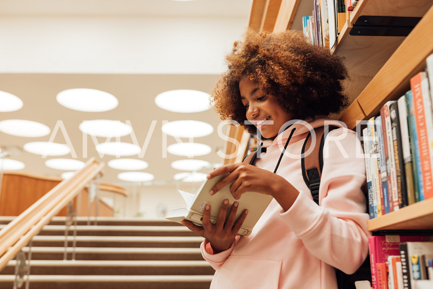 Smiling girl reading a book while leaning a bookshelf in library