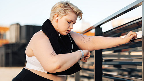 Side view of young plus-size woman looking at a smartwatch. Sweated and exhausted curvy woman with a towel around her neck relaxing during training.