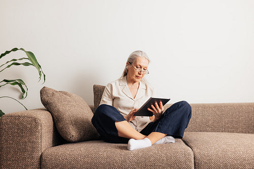 Senior woman sitting on a sofa with crossed legs holding digital tablet. Aged female reading from a portable computer at home.