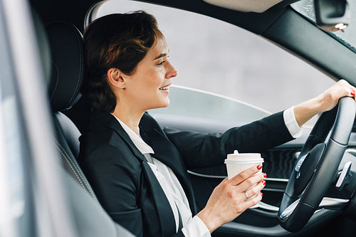 Side view of a smiling businesswoman holding a coffee and driving a car