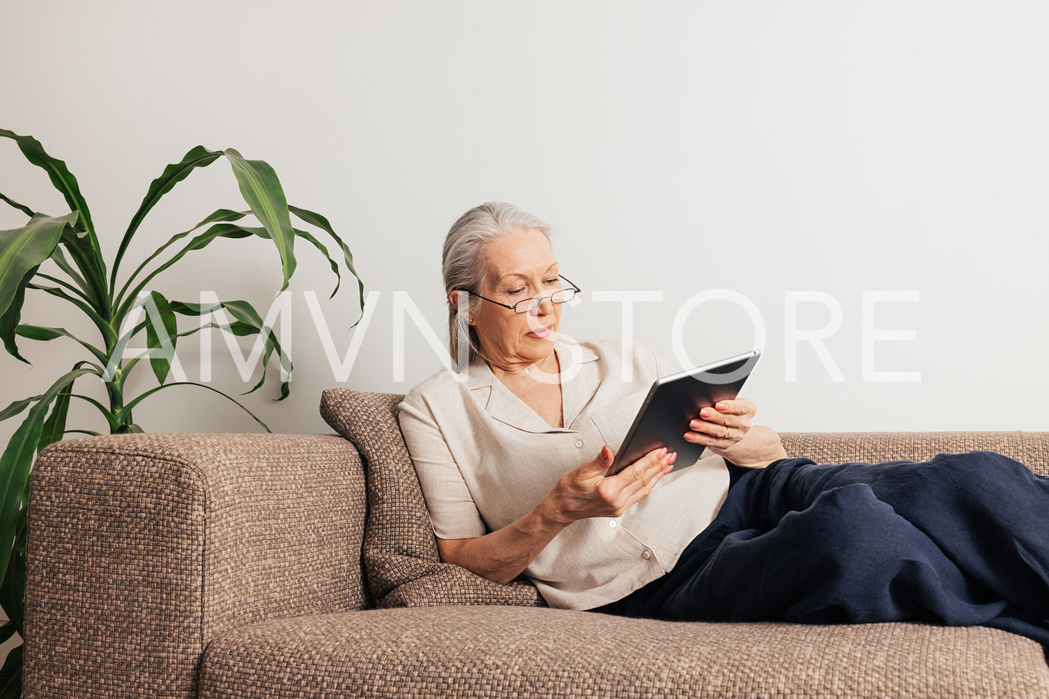 Senior woman in glasses lying on a sofa holding a digital tablet. Aged female in casuals reading from the portable computer.