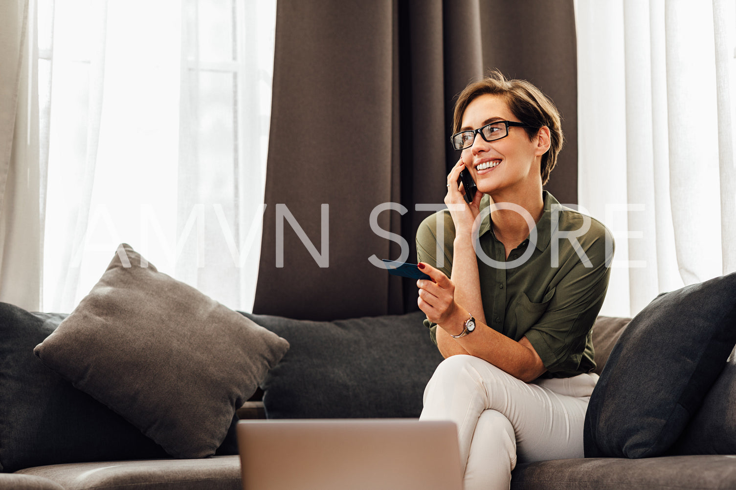 Beautiful smiling woman sitting on a couch in hotel room and talking on mobile phone. Young female entrepreneur in formal clothes looking away indoors.	