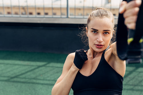 Young woman doing exercises on a rooftop. Fitness female working out outdoors using suspension strap for lifting.