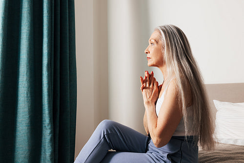 Side view of a senior woman with long hair looking thoughtful while sitting on a bed