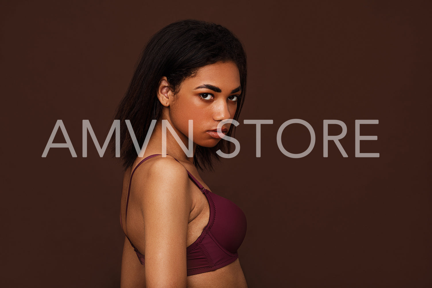 Side view of a young woman with black hair looking at camera. Young slim female in lingerie posing on brown background.