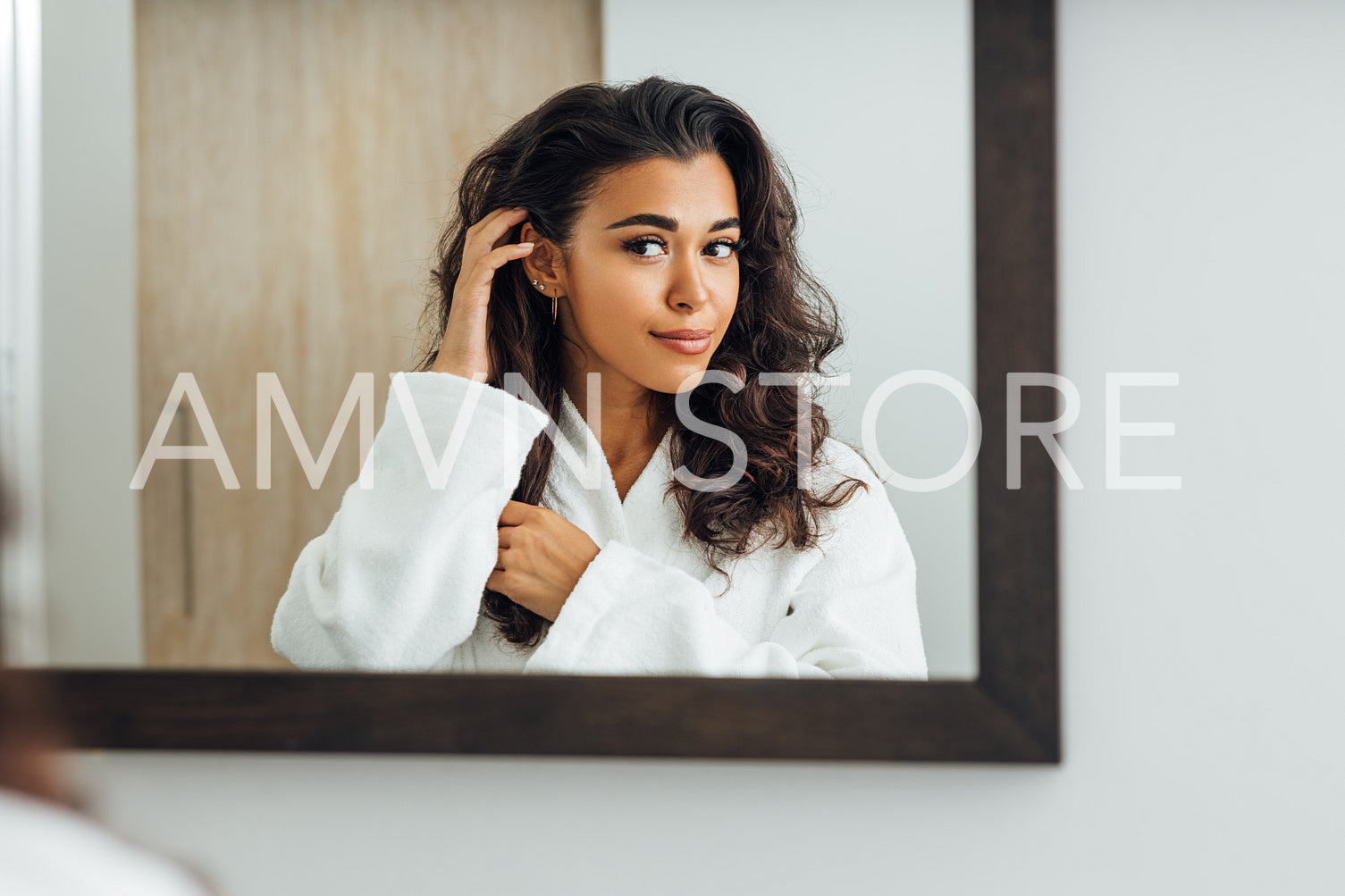 Beautiful female wearing bathrobe looking at a mirror and touching her hair	