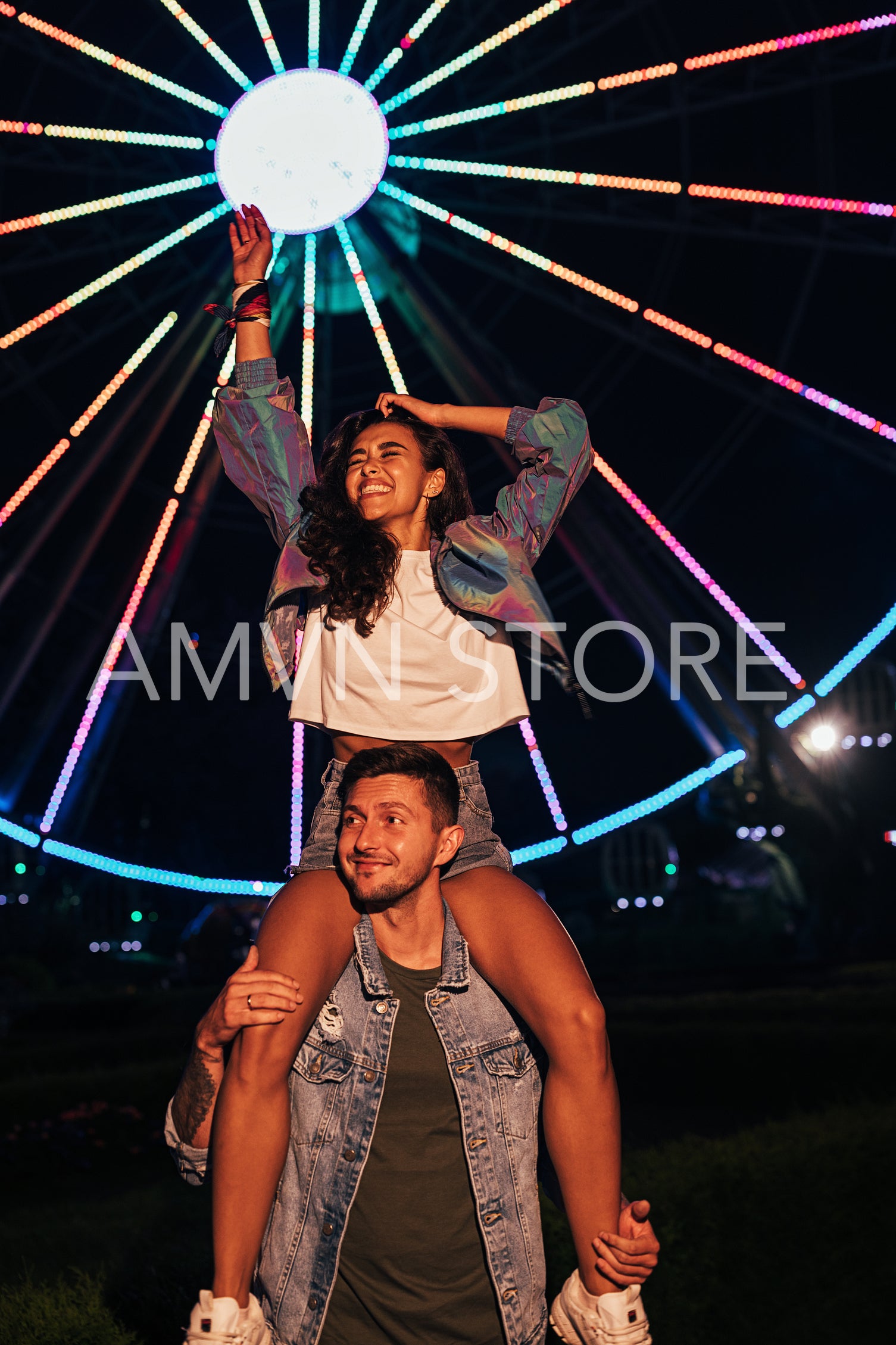 Young happy girlfriend sitting on the shoulders of her boyfriend and raising hand up in the amusement park