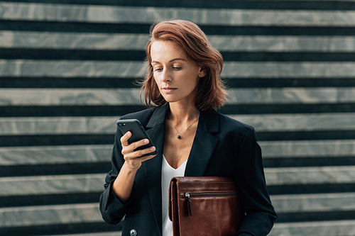 Confident middle-aged female with a leather folder looking at her smartphone while standing outdoors