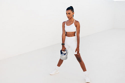 Woman standing with kettlebell outdoors. Female athlete exercising with weight in white outdoor studio.