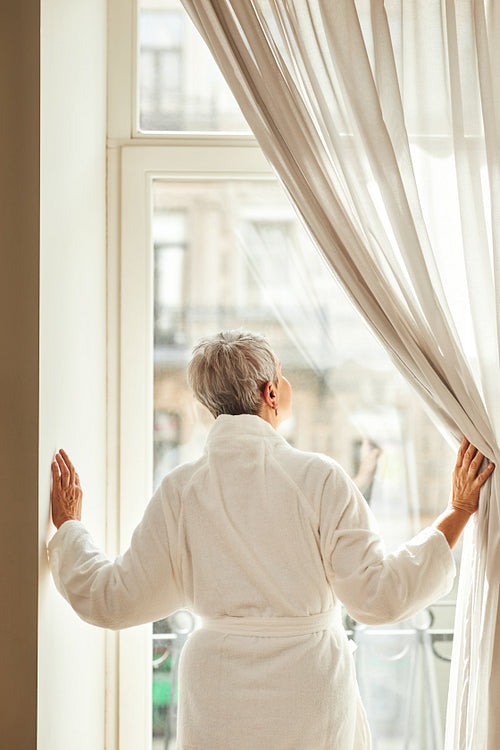 Rear view of a senior woman in bathrobe opening curtains in the morning