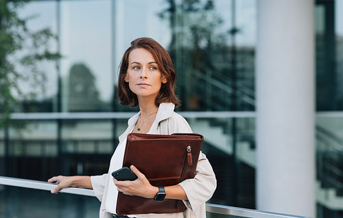 Confident female in coat with leather folder looking away while standing at railing against business building