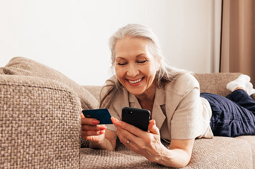 Close up of a senior female holding a credit card and smartphone. Adult smiling woman making a purchase with smartphone at home.