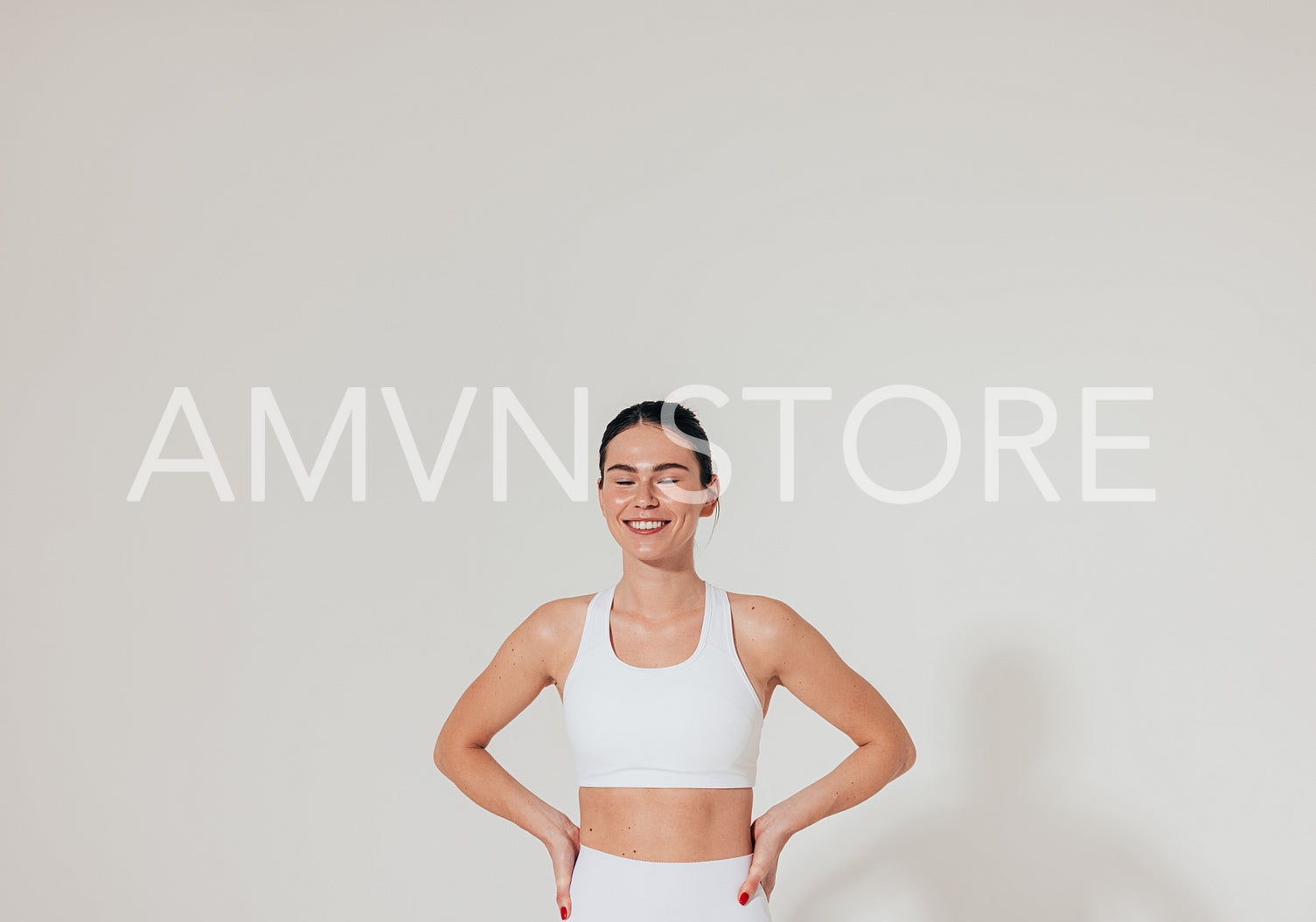 Smiling woman with closed eyes and hands on hips wearing white fitness attire standing at white wall