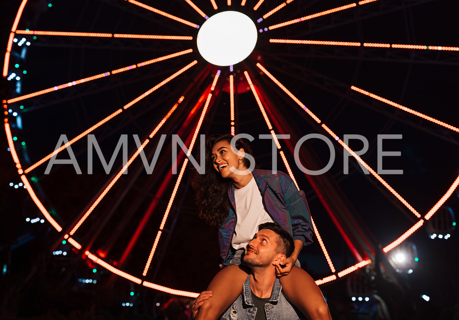 Young laughing female sitting on the shoulders of her boyfriend and looking away at night in an amusement park