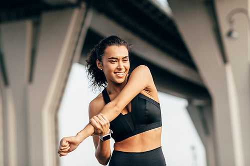 Woman athlete warming up her body before evening training under a highway. Smiling female stretching arm outdoors.