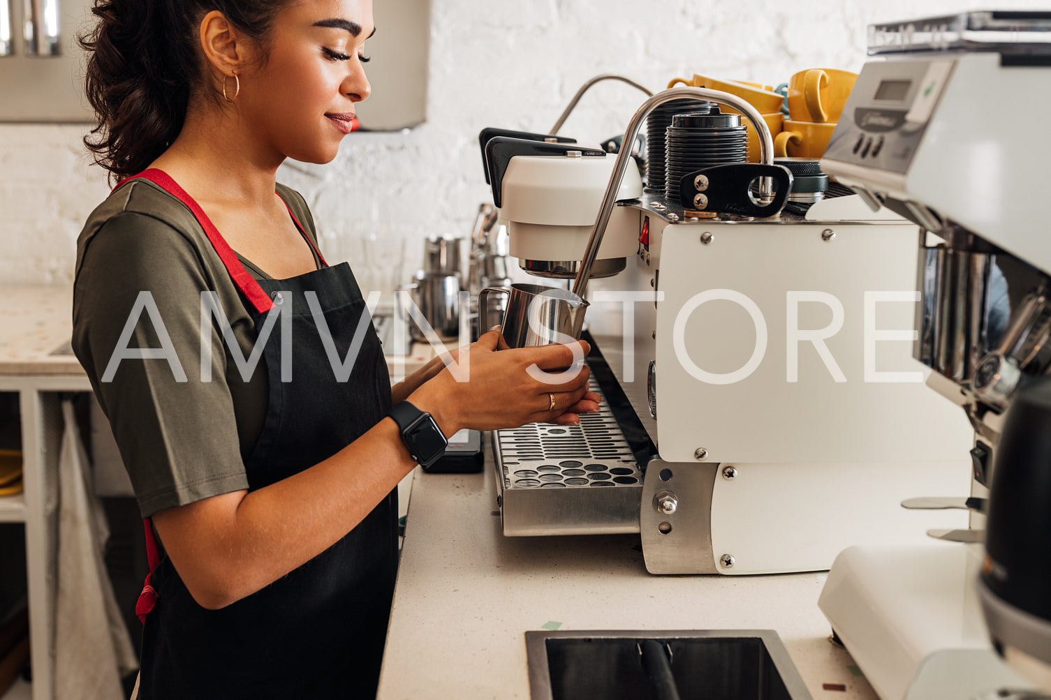 Side view of a female barista in apron standing at coffee machine and holding frothing pitcher	